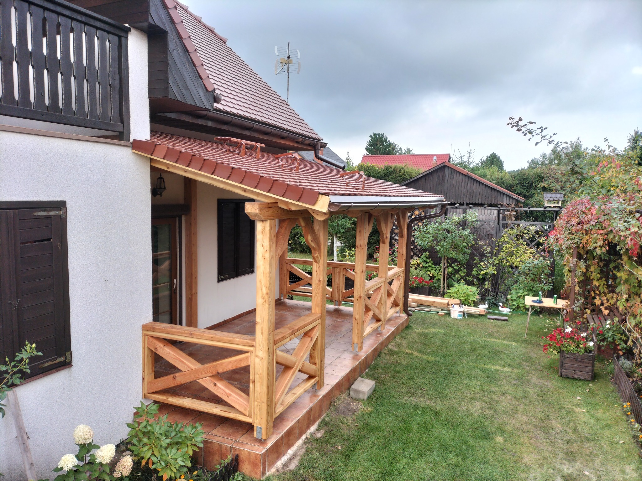 Zadaszony, drewniany taras przylegający do domu z białymi ścianami, widok z ogrodu, z elementami drewnianej konstrukcji w stylu rustykalnym i brązową terakotą na podłodze.