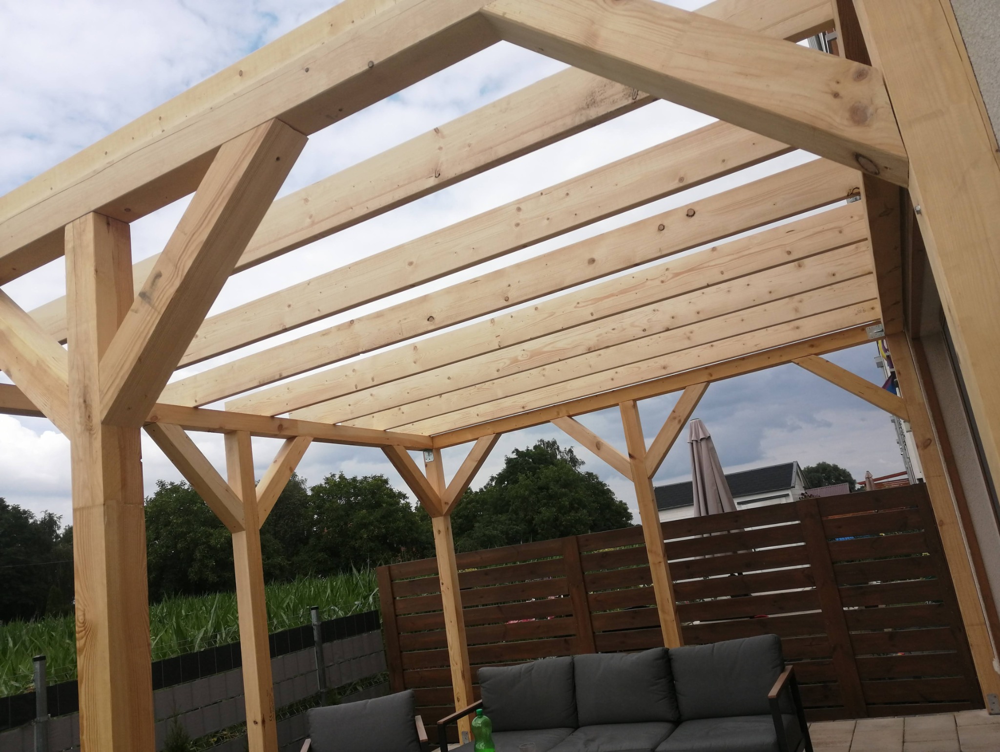 Surowa drewniana konstrukcja z belek, tworząca szkielet zadaszenia nad tarasem, widok od dołu, z widocznymi elementami mocującymi i fragmentem mebli ogrodowych.