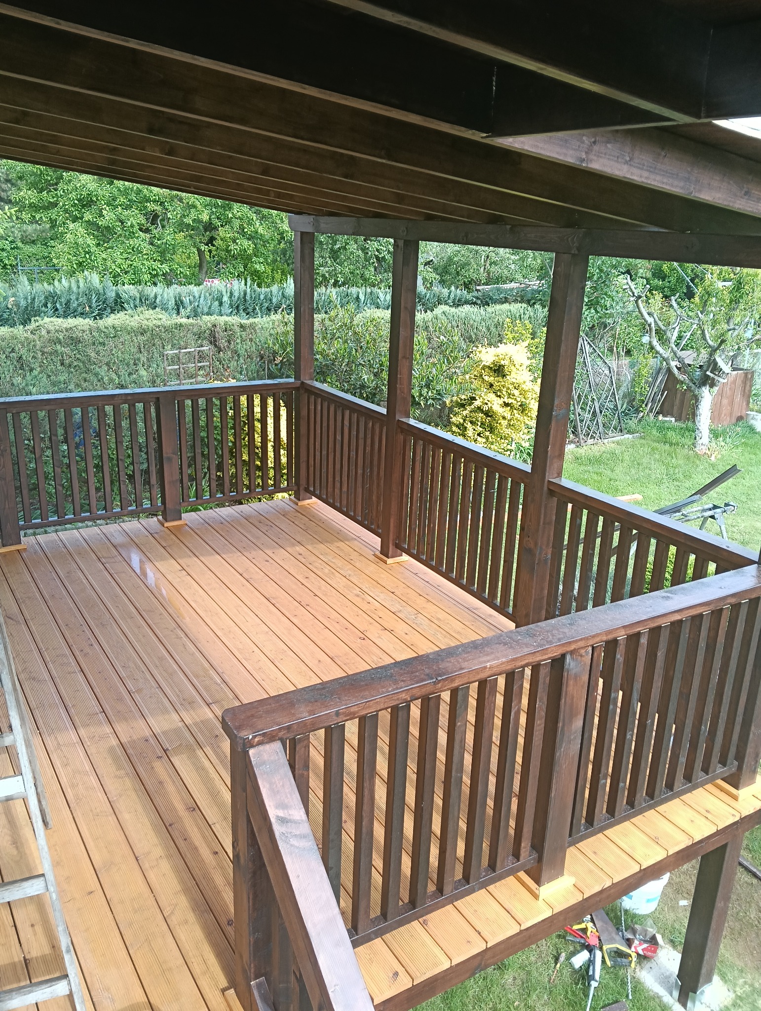 Zadaszony taras drewniany z balustradą, świeżo po impregnacji. Widoczne narzędzia pracy, otoczenie zieleni. Ujęcie z góry, ukazuje detale konstrukcji i wykończenia.
