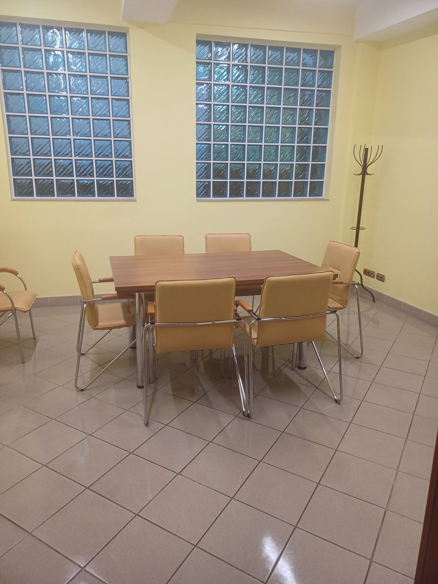 Sala konferencyjna z drewnianym stołem i sześcioma krzesłami obitymi skórą w odcieniach beżu, oświetlona przez dwa okna z luksferami i lampę-wieszak na ubrania