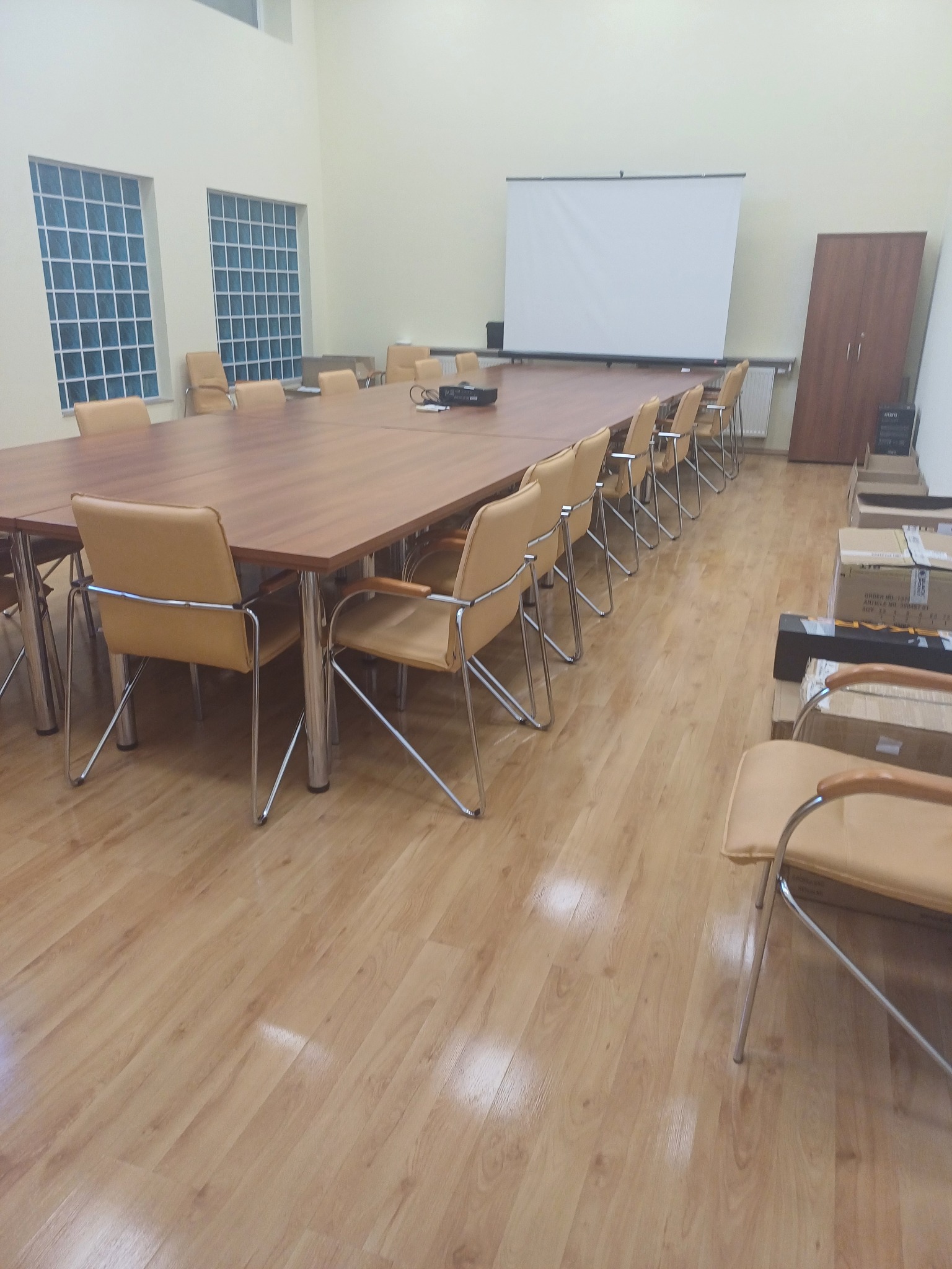 Sala konferencyjna z długim stołem, krzesłami z beżowej skóry i metalowymi nogami, rzutnikiem na stole i ekranem projekcyjnym, kartonami w rogu pomieszczenia, jasna podłoga.