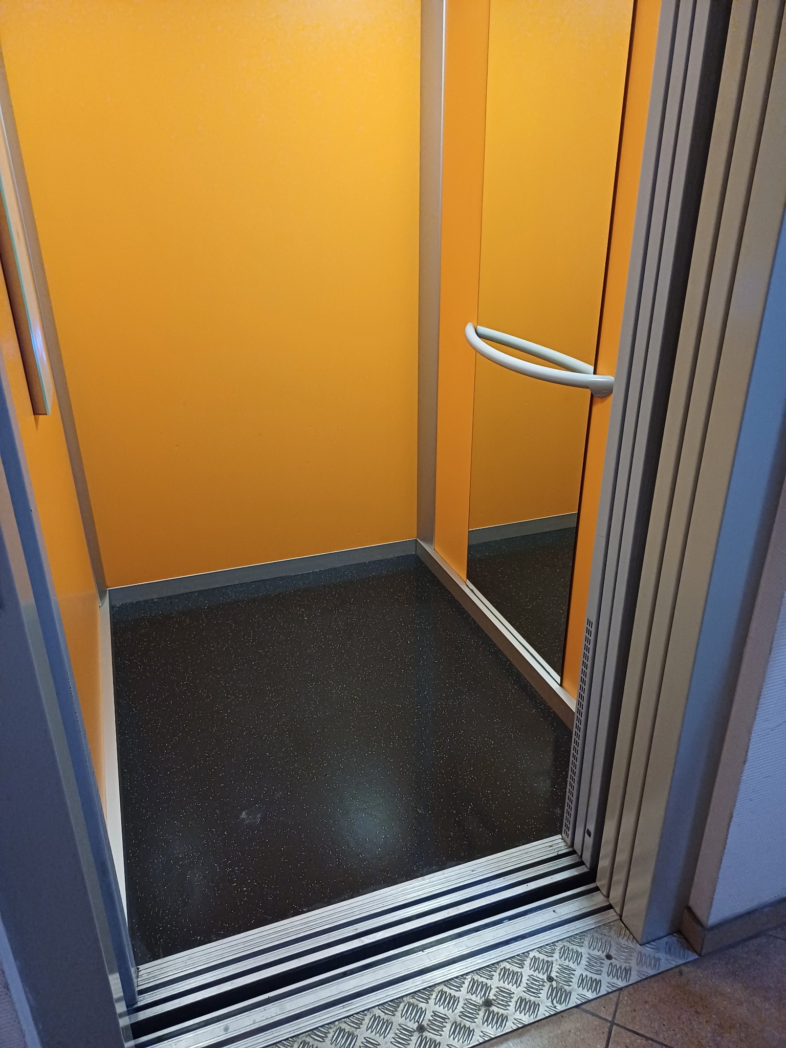 Wnętrze pustej, pomarańczowej kabiny windy z czarną podłogą i metalowymi drzwiami harmonijkowymi.