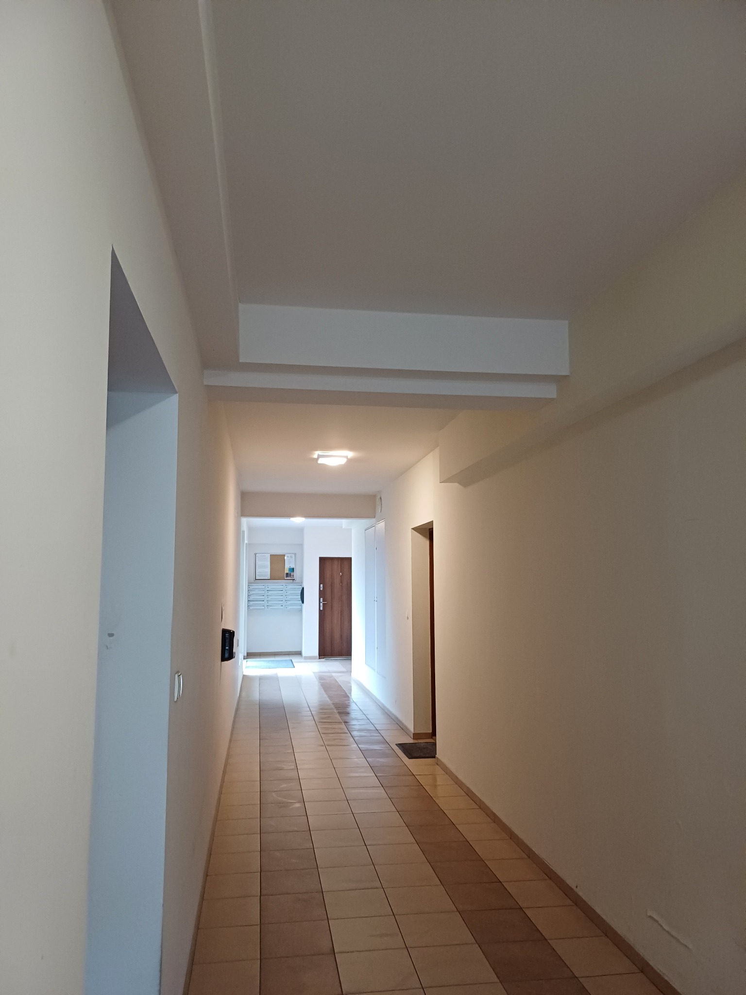 Długi korytarz w bloku mieszkalnym z beżowymi ścianami, brązowymi płytkami na podłodze i białym sufitem z wystającym elementem dekoracyjnym.