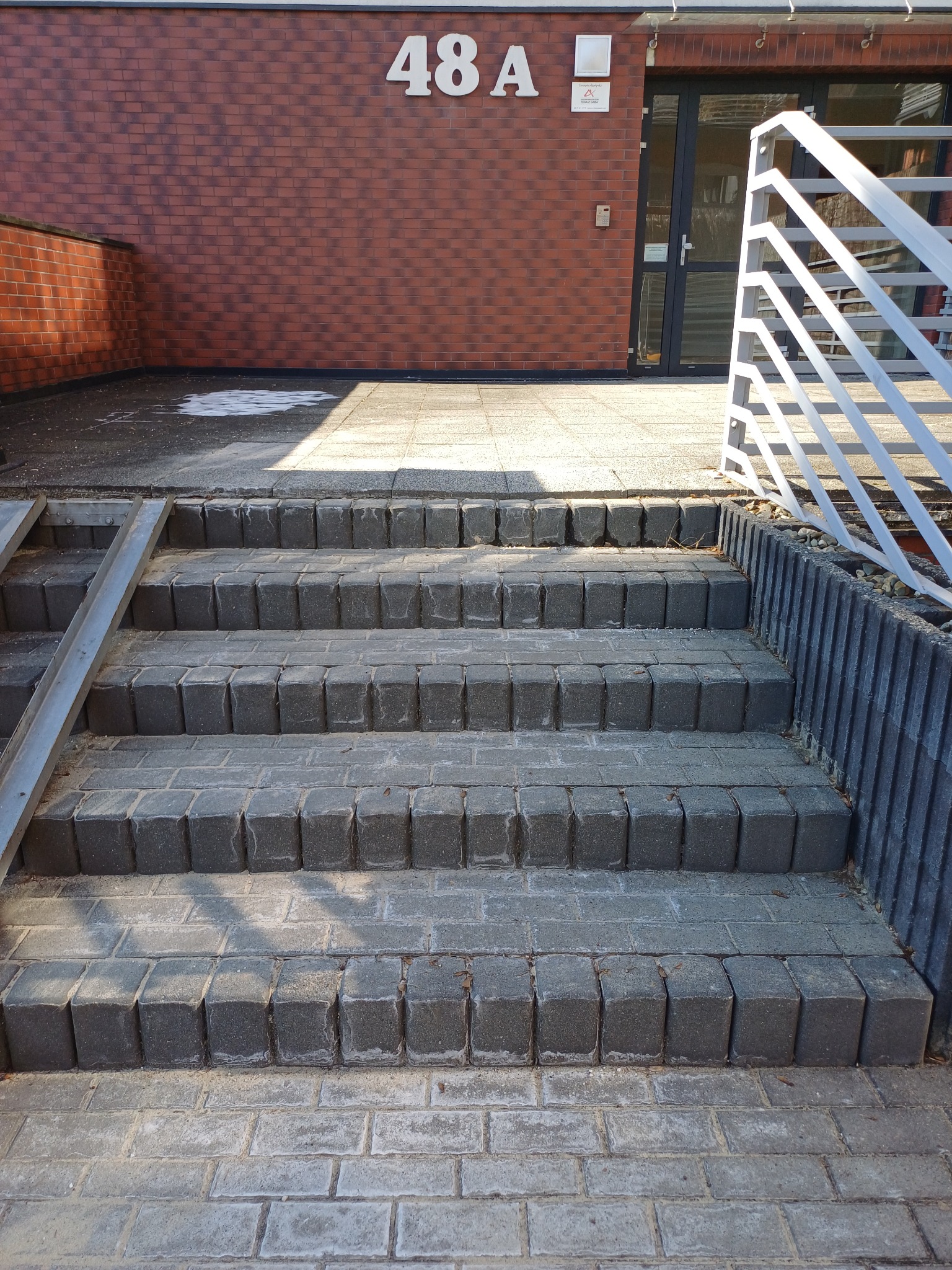 Schody z kostki brukowej prowadzące do budynku z numerem 48A, widoczna barierka i fragment rampy dla wózków.