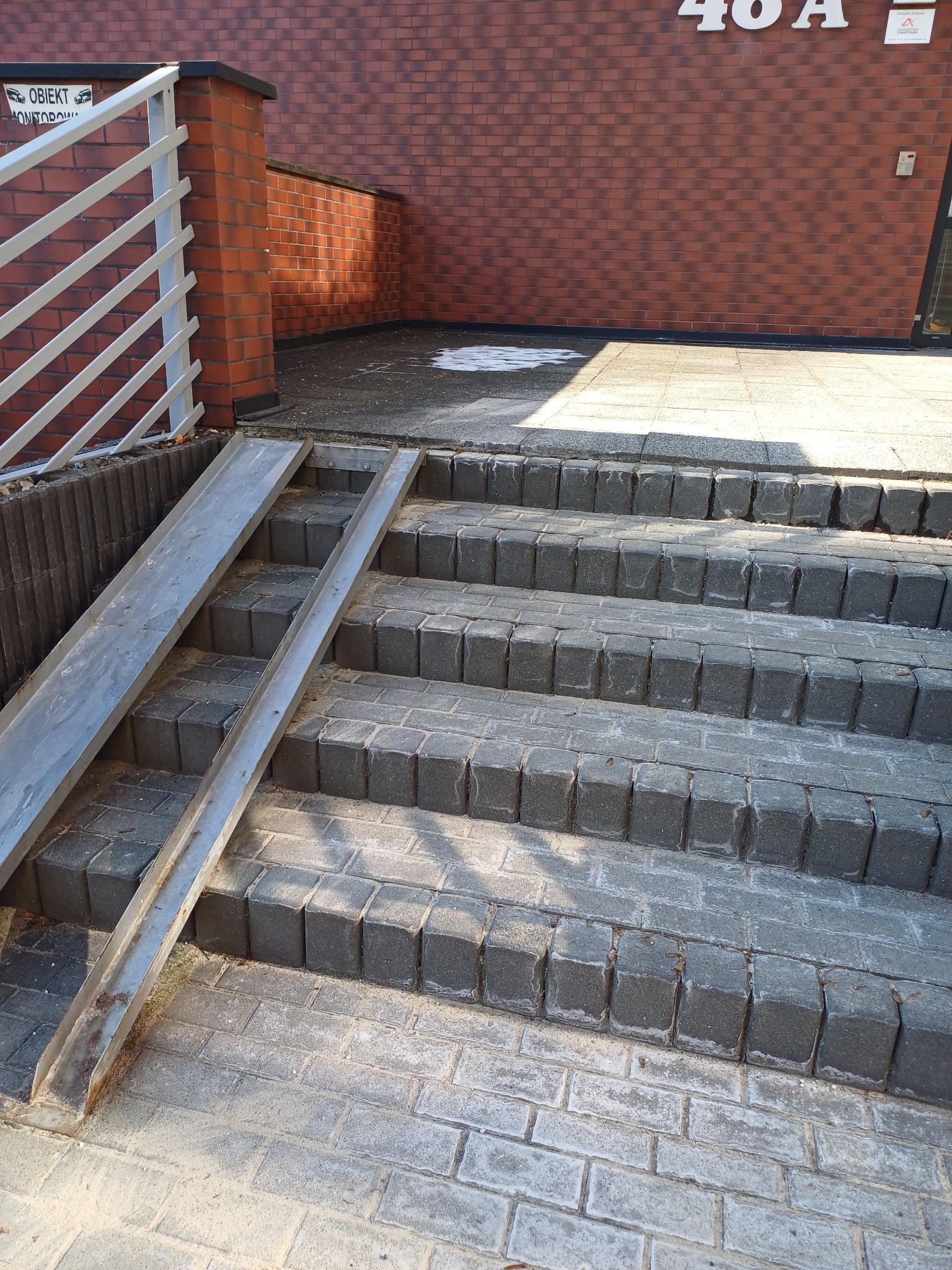 Zewnętrzne schody z szarej kostki brukowej z metalową rampą dla wózków inwalidzkich, prowadzące do budynku z ceglaną elewacją, widoczne ślady zabrudzeń na kostce.