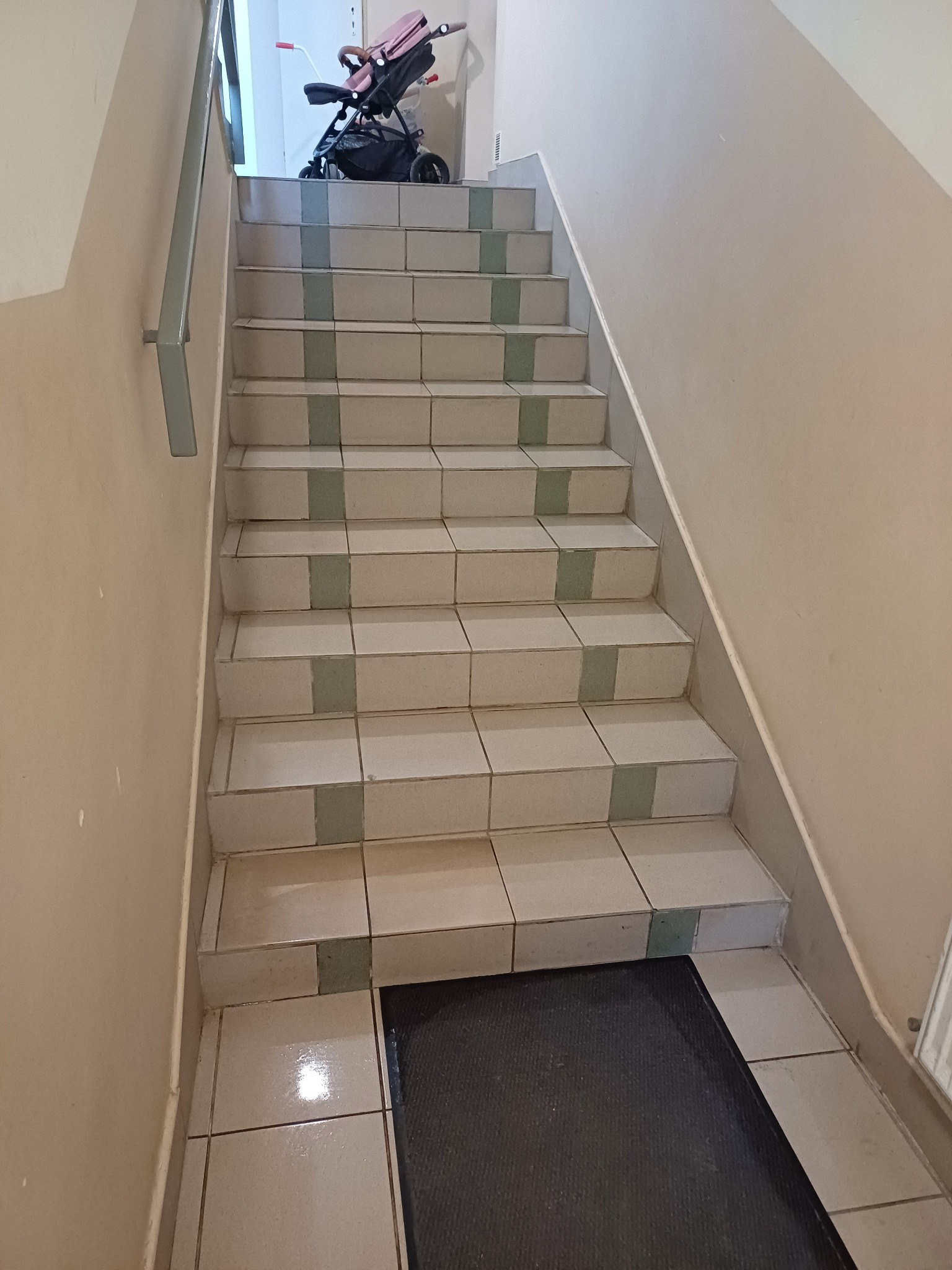 Klatka schodowa z wyłożonymi płytkami schodami, na górze widoczny wózek dziecięcy, a na dole wycieraczka.