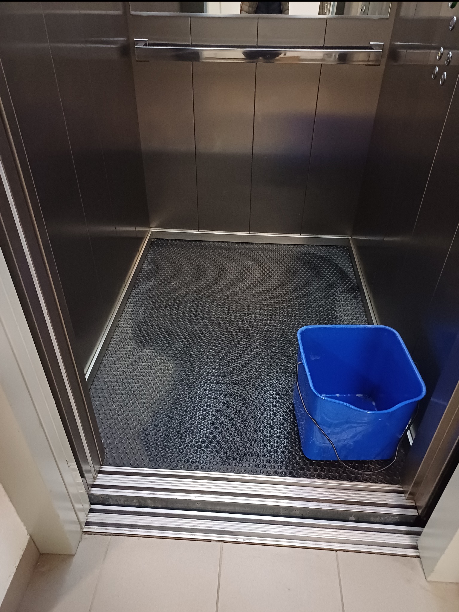 Wnętrze windy w Wilkowicach podczas sprzątania, widoczne wiadro z wodą i czyszczona podłoga z fakturą.