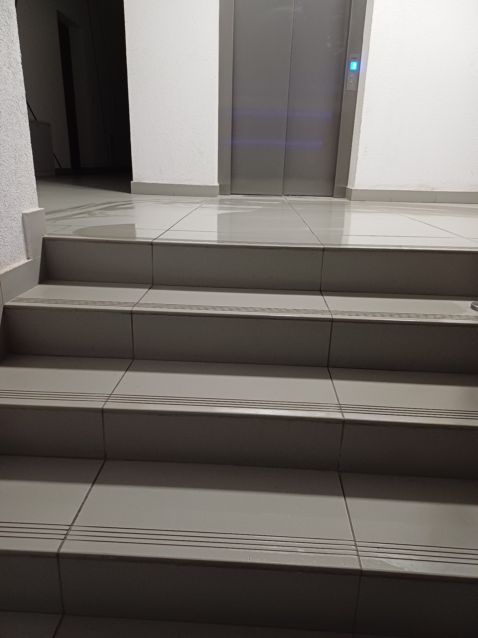 Klatka schodowa z szarymi płytkami i metalowymi schodami prowadząca do windy z niebieskim podświetleniem przycisku.