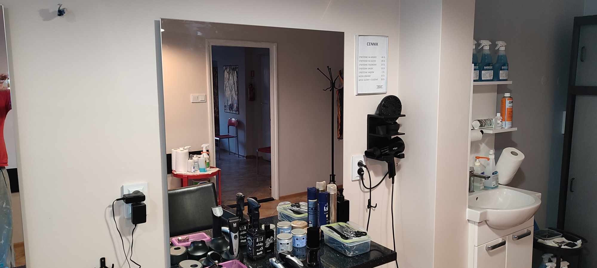 Wnętrze salonu fryzjerskiego z dużym lustrem, na blacie widoczne narzędzia i kosmetyki fryzjerskie, na ścianie cennik usług, w tle widoczny fotel fryzjerski i dodatkowe wyposażenie.