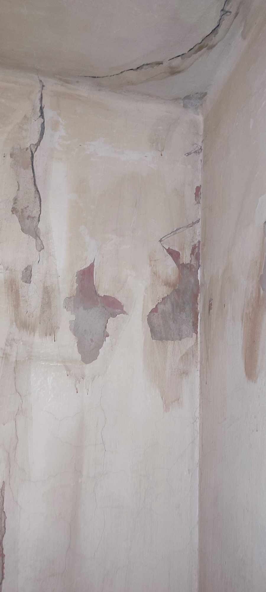 Narożnik pomieszczenia z odpadającym tynkiem, widoczne liczne pęknięcia i ślady wilgoci, fragmenty starej farby w kolorze czerwonym.