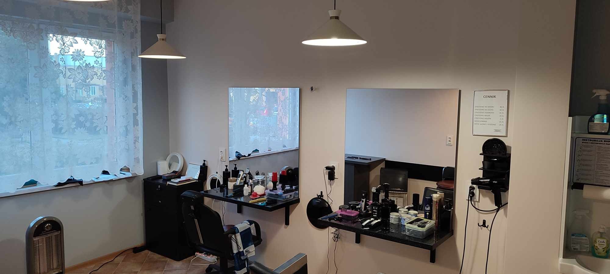 Wnętrze salonu fryzjerskiego z dwoma stanowiskami, lustrami, czarnymi półkami z kosmetykami i narzędziami, cennikiem na ścianie oraz lampami sufitowymi.
