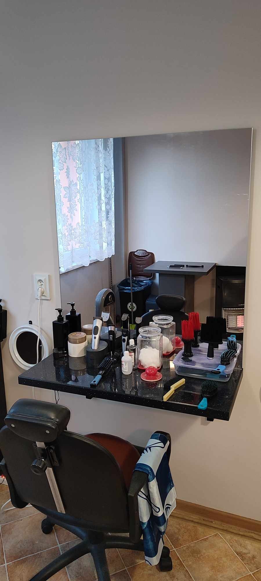 Stanowisko fryzjerskie z czarnym blatem, lustrem i różnymi akcesoriami, w tle okno z białą firanką.