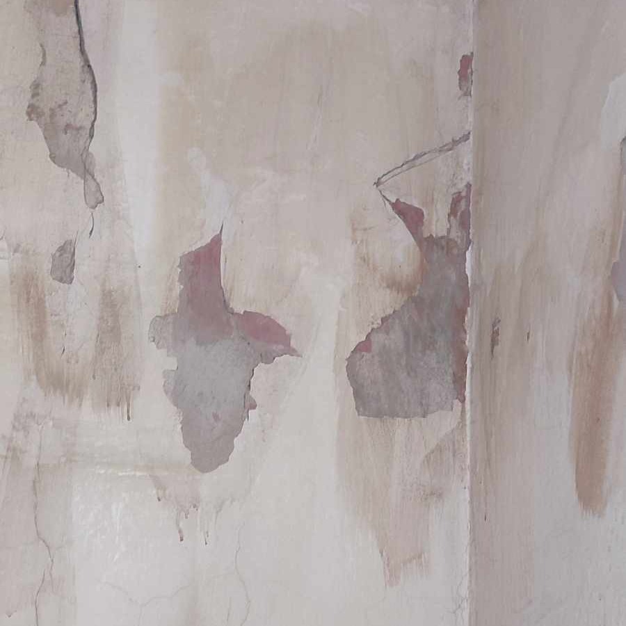Zniszczona ściana w rogu pomieszczenia z odpadającą farbą, odsłaniająca warstwy poprzednich malowań w odcieniach różu i szarości.