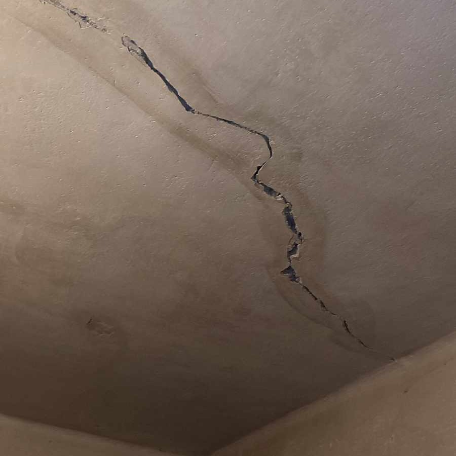 Pęknięcie na suficie z widocznymi śladami wilgoci, biegnące przez całą powierzchnię, z perspektywy narożnika pokoju.