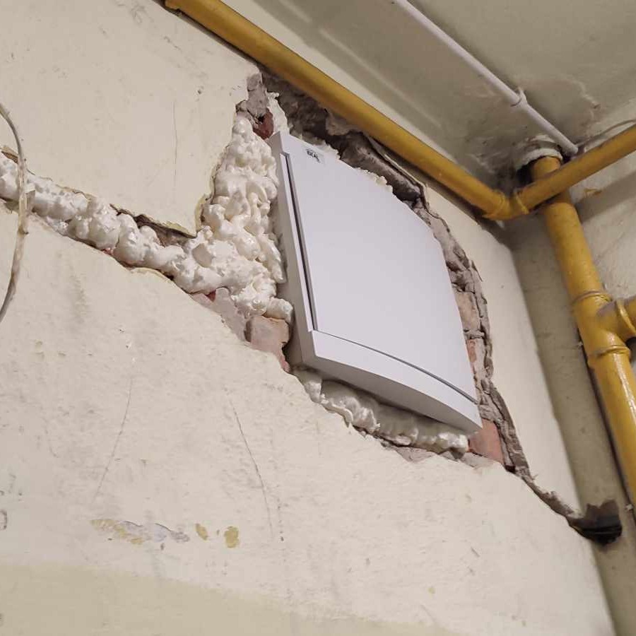 Biała skrzynka elektryczna zamontowana w ścianie z widocznymi ubytkami wypełnionymi pianką montażową, żółte rury gazowe w tle.