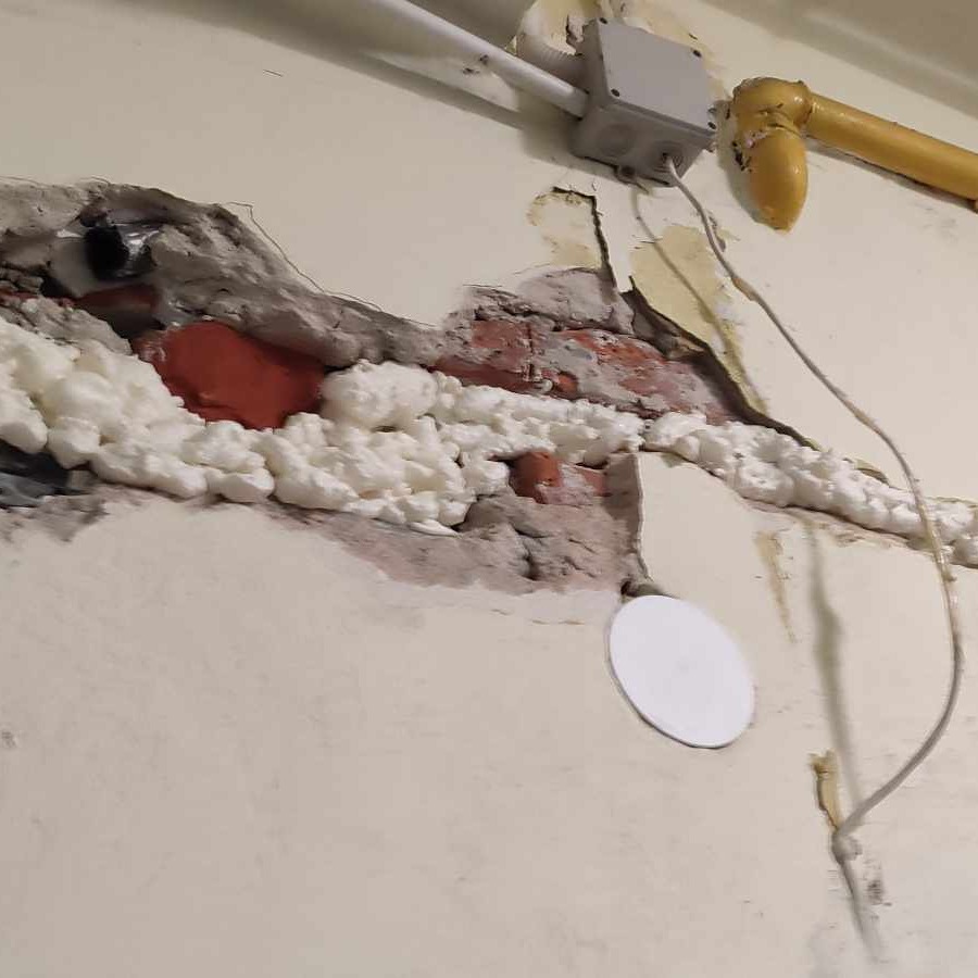 Odsłonięta ściana z fragmentami cegieł, wypełniona pianką montażową i zaprawą, z widocznymi przewodami elektrycznymi i rurami.