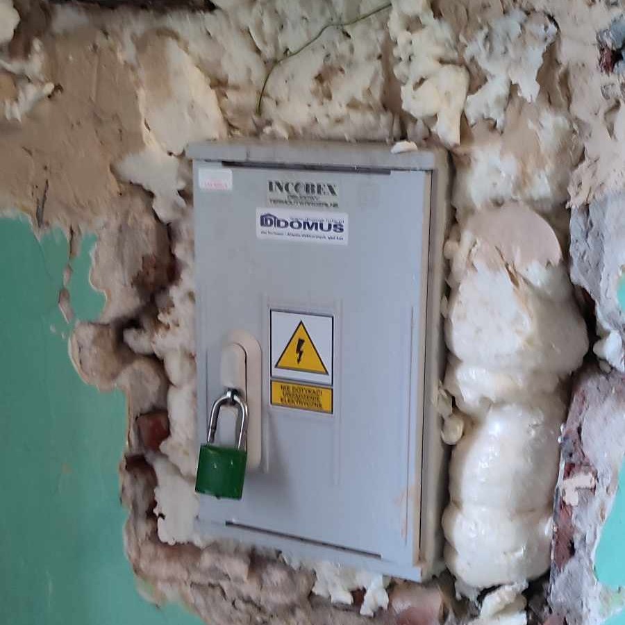 Szafka elektryczna z zieloną kłódką, otoczona pianką montażową, na tle fragmentu zielonej ściany z widocznymi ubytkami tynku.