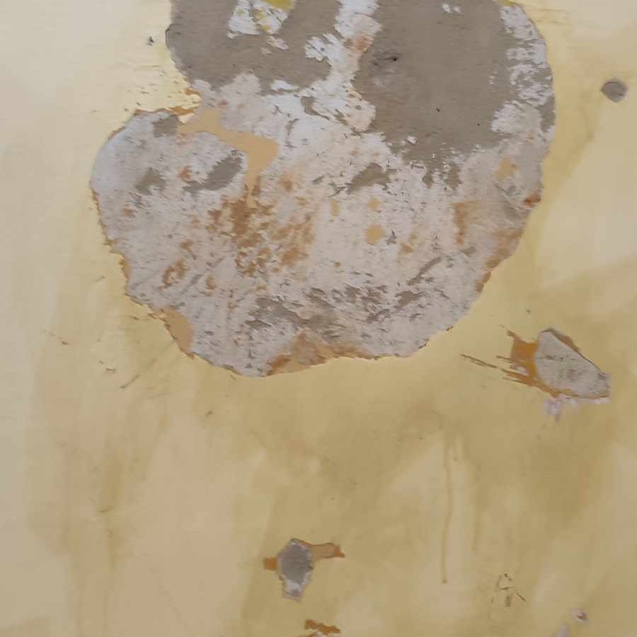 Żółta ściana z dużym, nieregularnym ubytkiem tynku, odsłaniającym szarą warstwę pod spodem i fragmenty wcześniejszej, żółtej warstwy farby.