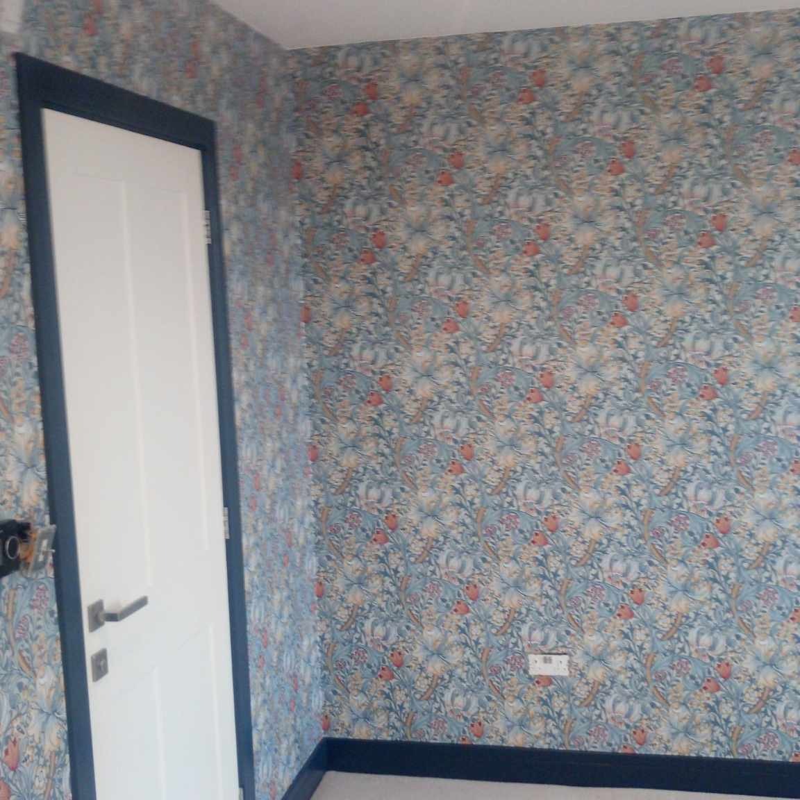 Pokój po remoncie: ściany pokryte tapetą w drobny, roślinny wzór w odcieniach niebieskiego, pomarańczowego i beżu, białe drzwi z granatową ościeżnicą i listwą przypodłogową.