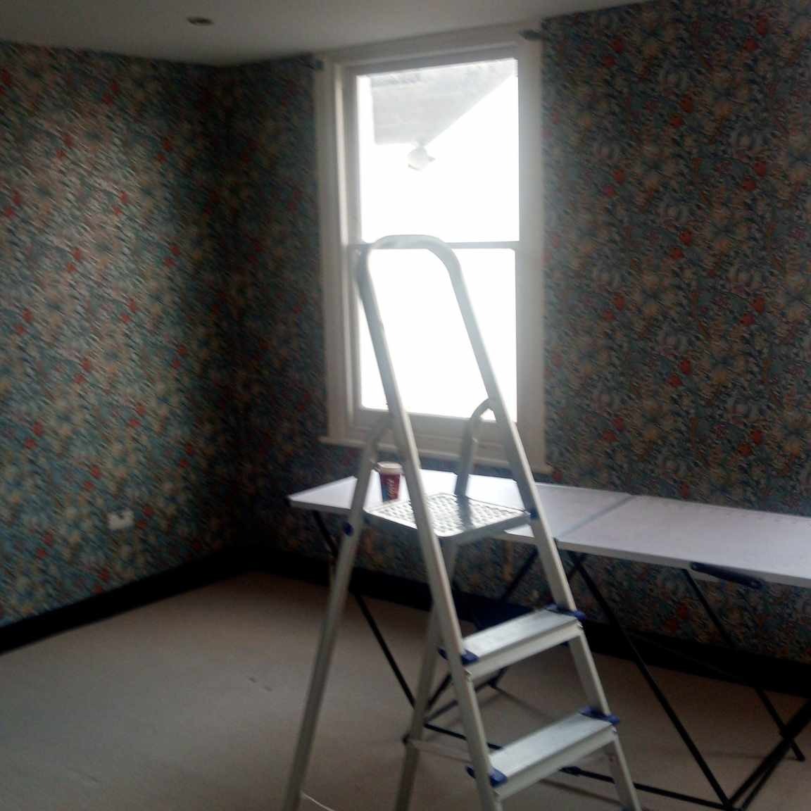 Pokój z tapetą w kwiaty, drabina aluminiowa i stół składany ustawione przy oknie, kubek na drabinie.
