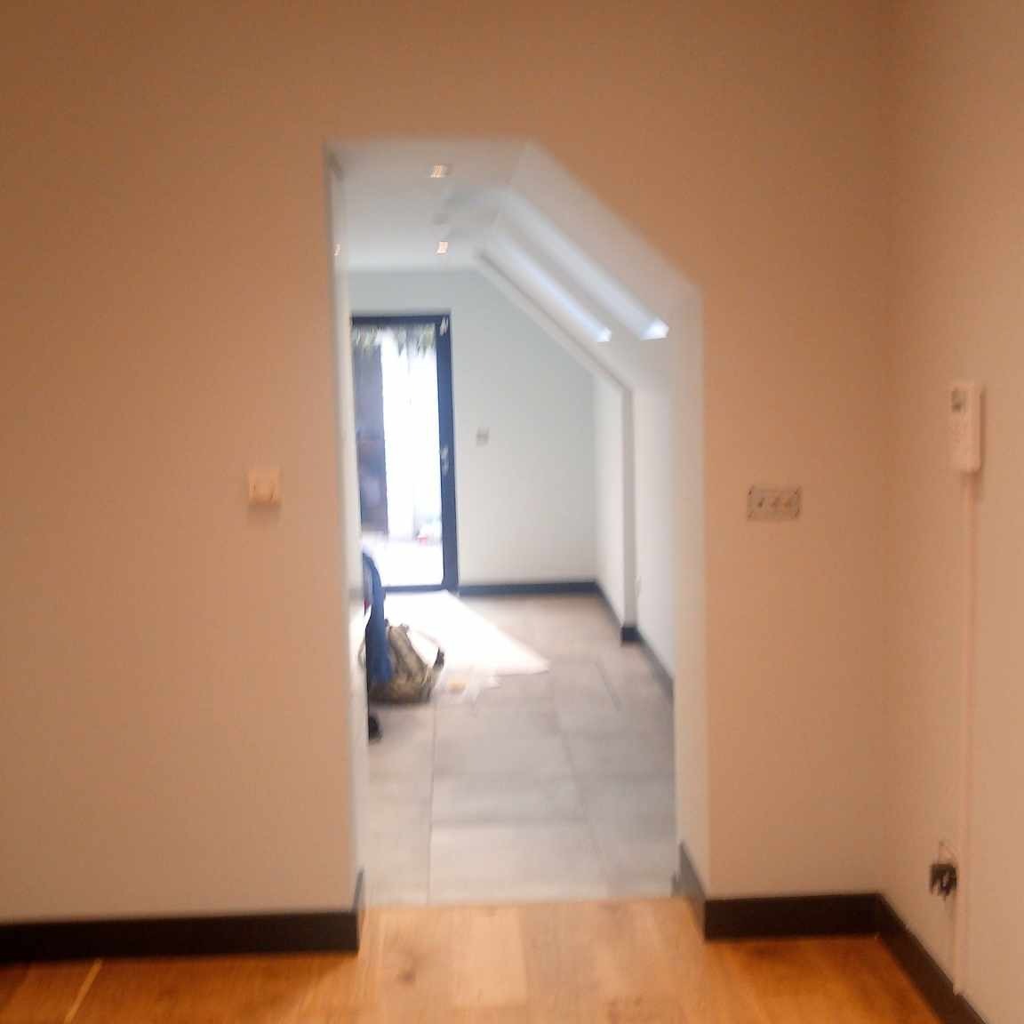 Wnętrze po remoncie z odmalowanymi ścianami w odcieniach beżu i bieli, widoczne gniazdka elektryczne, drewniana podłoga i fragment korytarza z szarą podłogą oraz otwartymi drzwiami wejściowymi w tle.