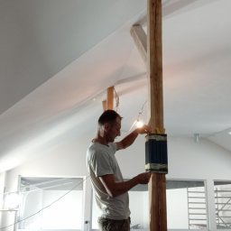 Reno bud - Pracownik przy drewnianej kolumnie w jasnym wnętrzu, prawdopodobnie podczas prac wykończeniowych. Widoczne narzędzia i elementy konstrukcyjne.