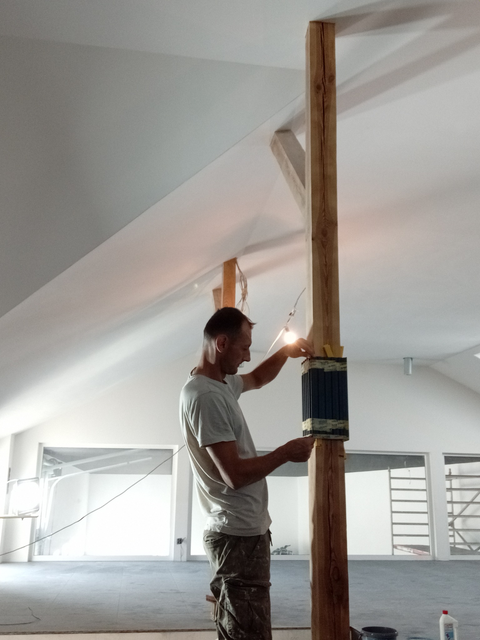 Pracownik przy drewnianej kolumnie w jasnym wnętrzu, prawdopodobnie podczas prac wykończeniowych. Widoczne narzędzia i elementy konstrukcyjne.