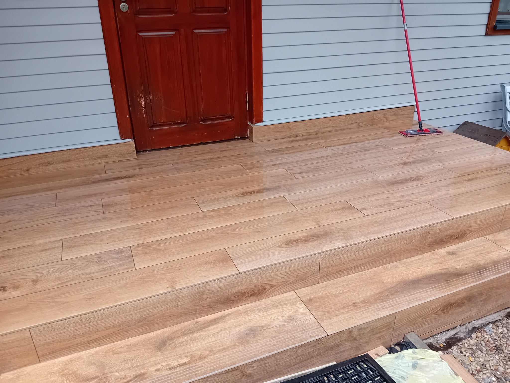 Wykończone schody wejściowe z płytek imitujących drewno, prowadzące do brązowych drzwi. Ściana budynku z jasnymi panelami. Widoczny mop do czyszczenia.