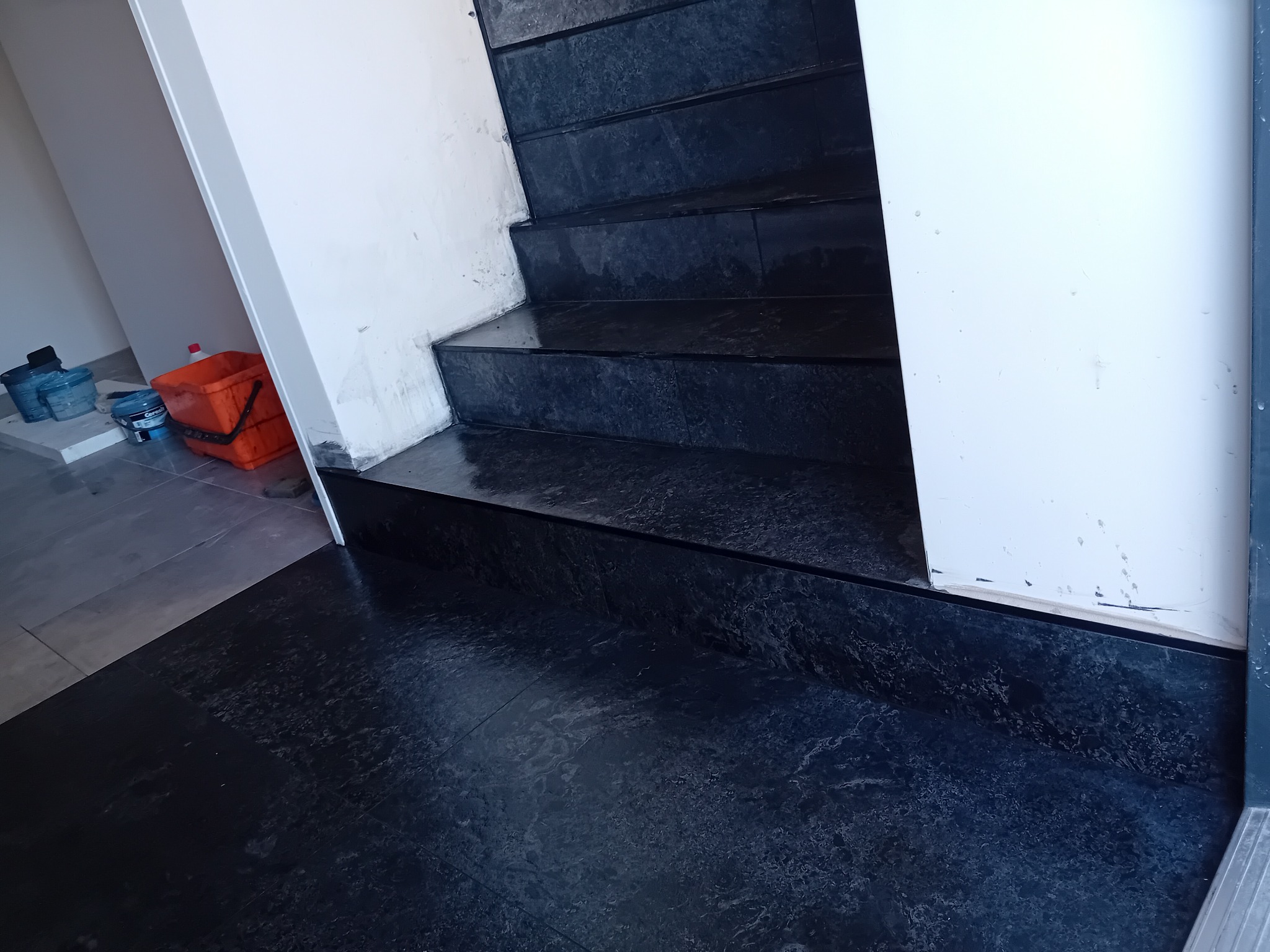 Wykończone, ciemne schody z płytek o wzorze marmuru, z widocznymi narzędziami i wiaderkami w tle. Ujęcie z dołu, podkreślające fakturę i detale wykończenia.