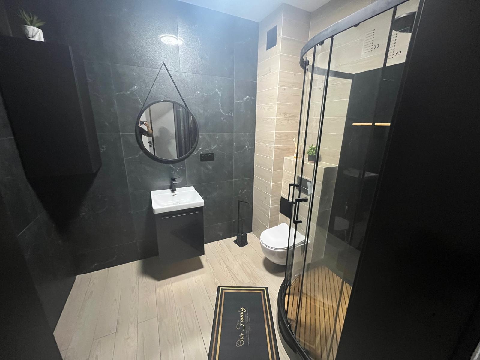 Nowoczesna łazienka z czarnymi płytkami imitującymi marmur, okrągłym lustrem na zawieszce, białą umywalką z szafką, kabiną prysznicową z czarnymi profilami i drewnopodobną podłogą oraz dywanikiem...