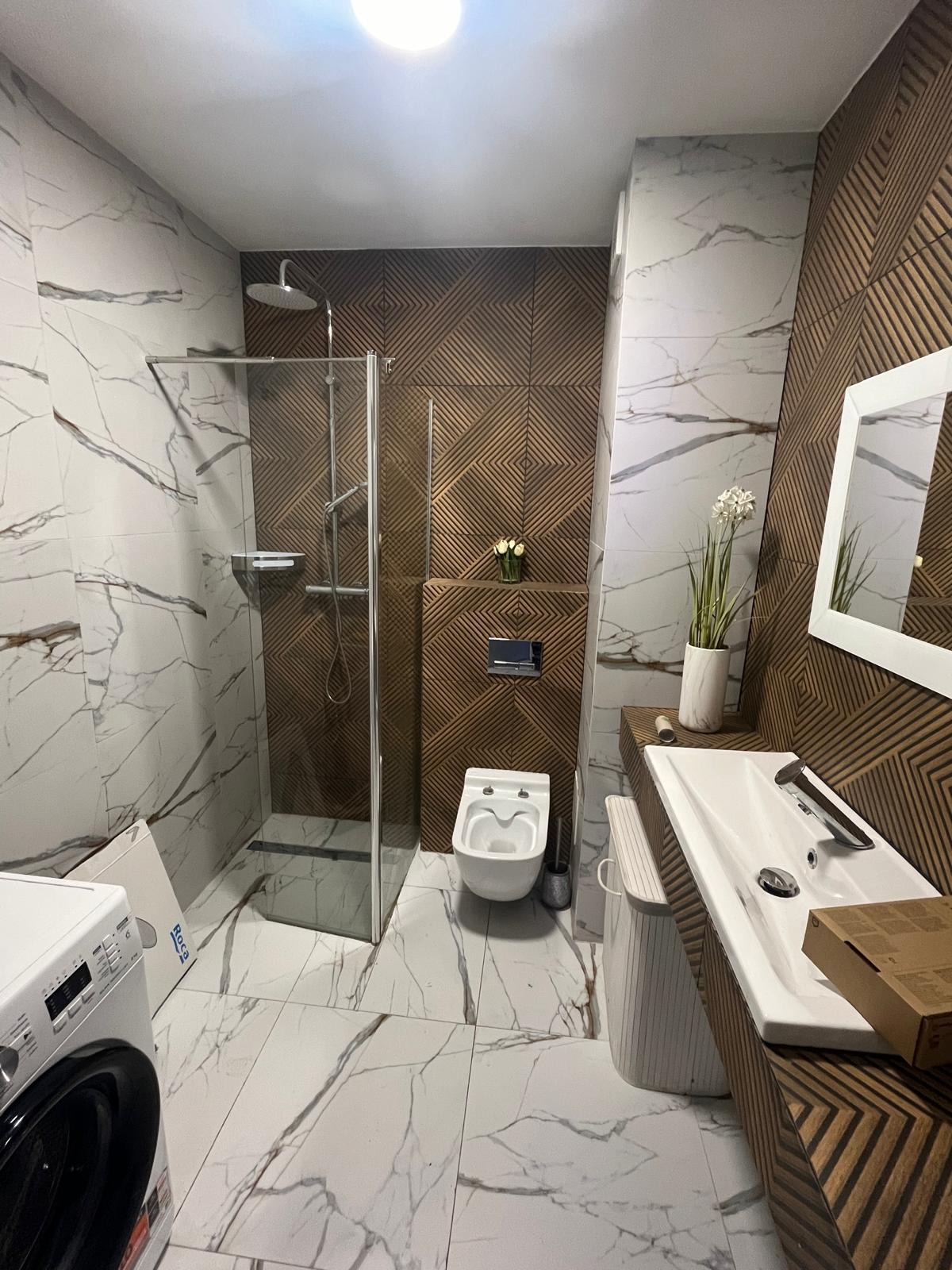 Nowoczesna łazienka z kabiną prysznicową, umywalką nablatową na drewnianym blacie z geometrycznym wzorem, toaletą podwieszaną i pralką Roca. Ściany wyłożone płytkami imitującymi marmur...