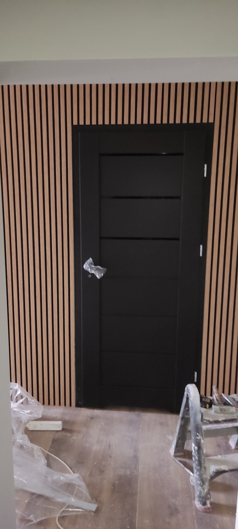 Czarna, nowoczesna stolarka drzwiowa z poziomymi panelami, zamontowana na ścianie z drewnianych listew, widoczne ślady prac wykończeniowych i drabina malarska.