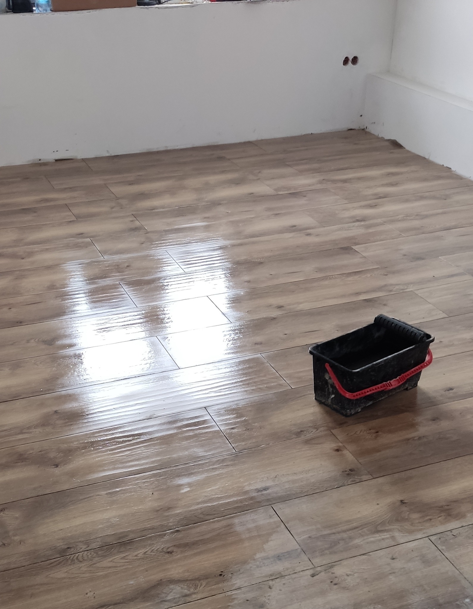 Świeżo położona, błyszcząca podłoga z paneli imitujących drewno, z widocznym czarnym, brudnym wiadrem z czerwoną rączką w rogu pomieszczenia i fragmentem białej ściany z gniazdkami.