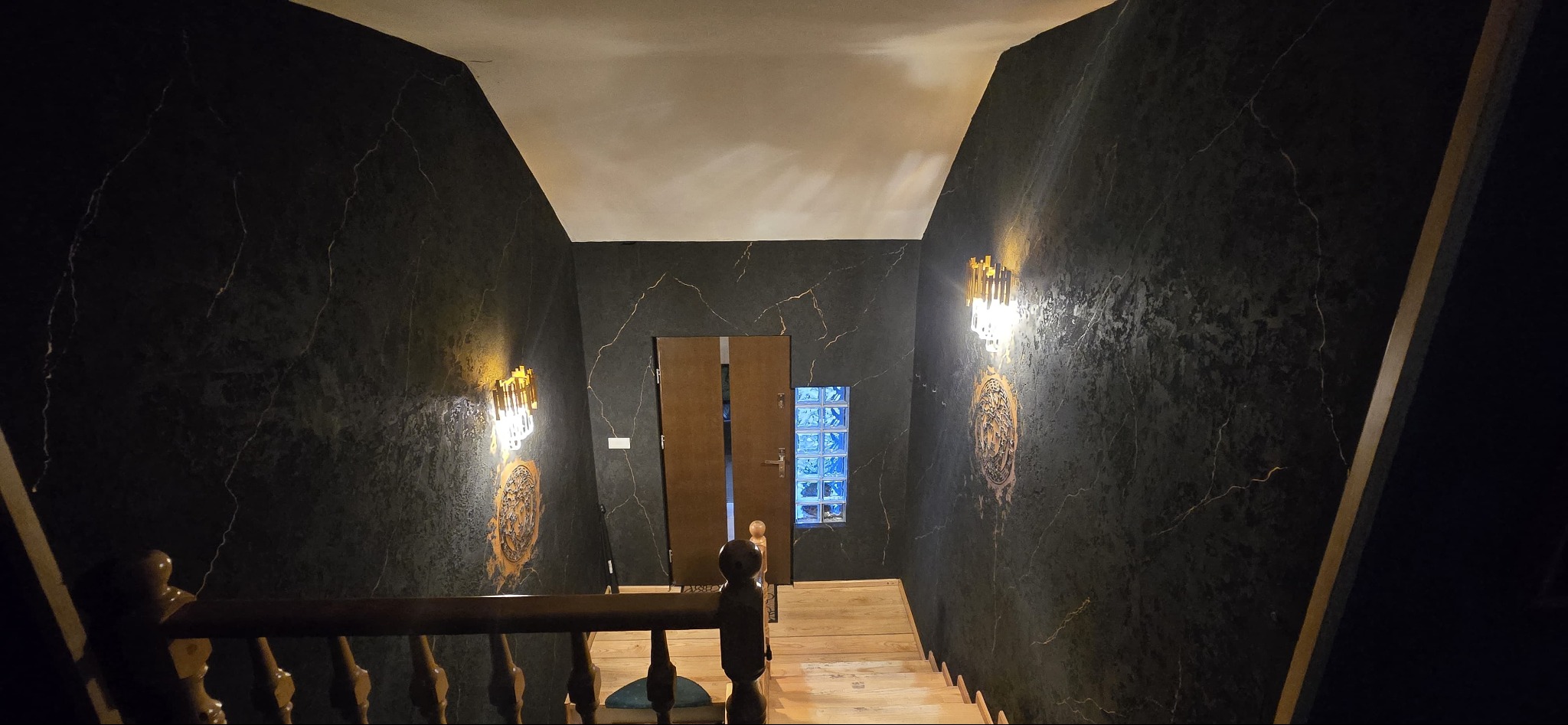 Klatka schodowa z czarnymi ścianami imitującymi marmur, oświetlona złotymi kinkietami i prowadząca do drewnianych drzwi z luksferami.