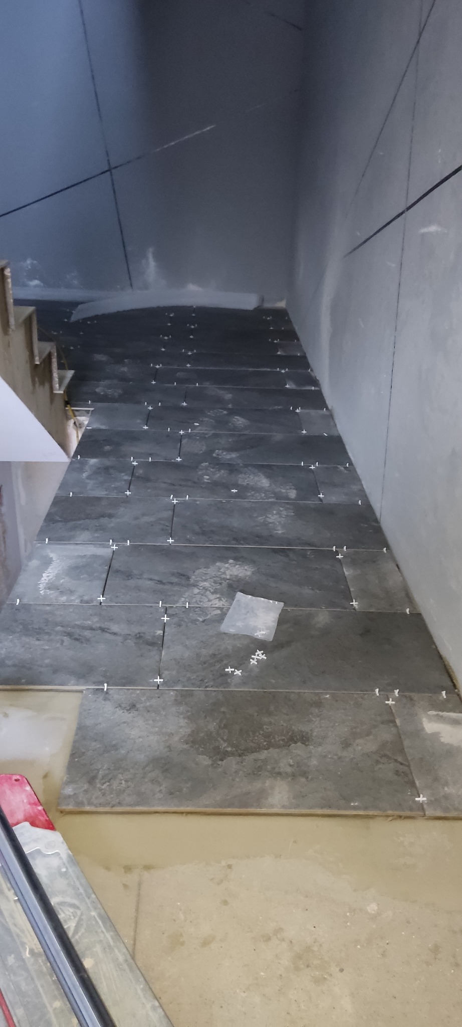 Układanie ciemnoszarych płytek na schodach z użyciem krzyżyków dystansowych, widoczne narzędzie do cięcia płytek w dolnym rogu.