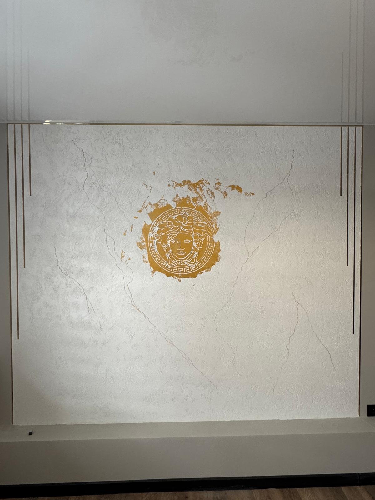 Ściana z dekoracyjną, marmurkową fakturą i złotym motywem Meduzy, obramowana pionowymi, złotymi liniami.