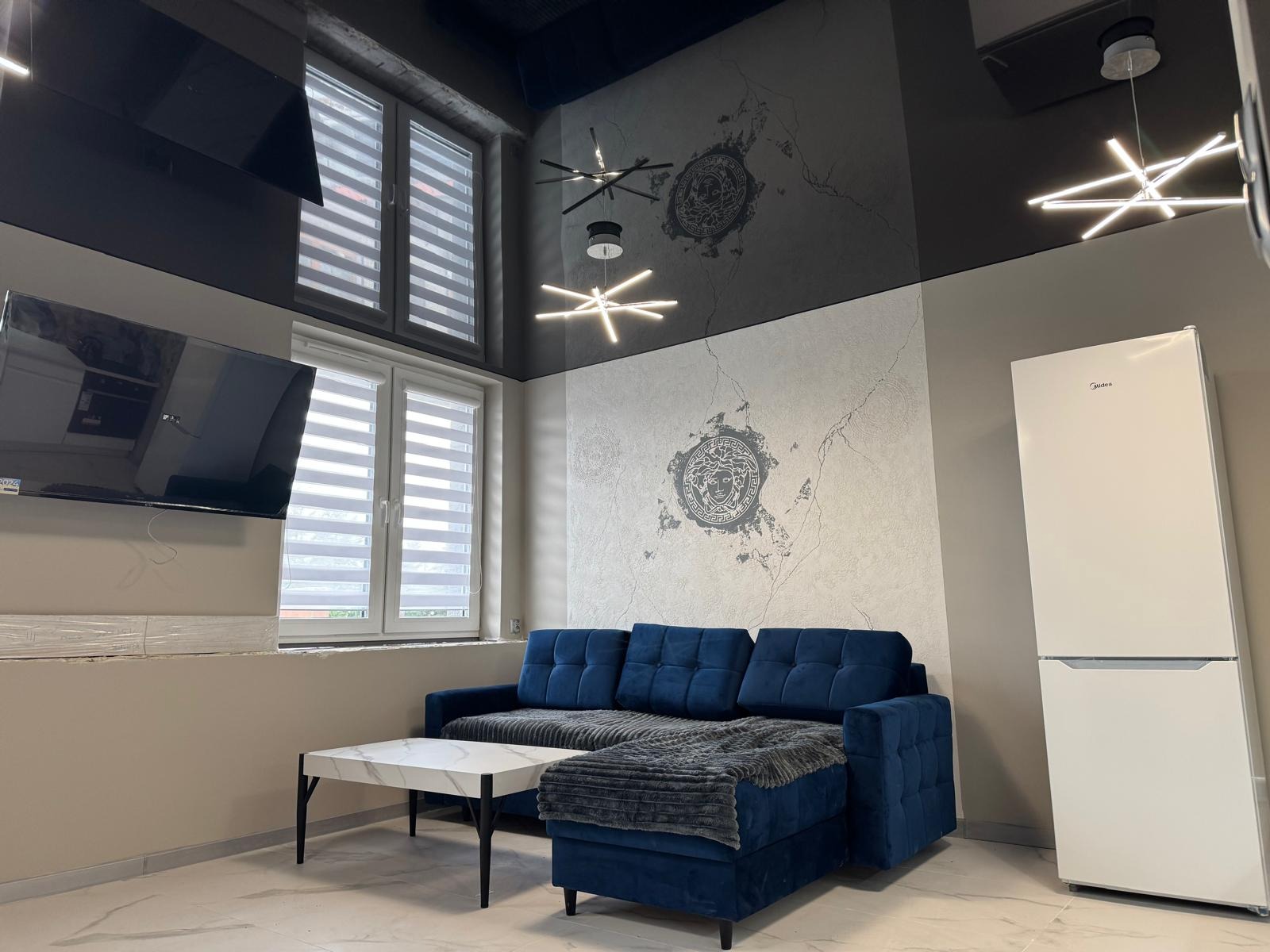 Salon z granatową sofą, marmurowym stolikiem i białą lodówką na tle ściany z motywem Versace, oświetlony nowoczesnymi lampami i doświetlony przez okna z żaluzjami.