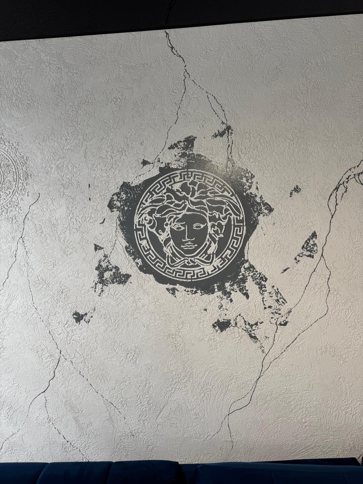 Dekoracyjna ściana z szarą, teksturowaną gładzią i motywem głowy Meduzy w stylu Versace, otoczonym imitacją spękań i uszkodzeń.