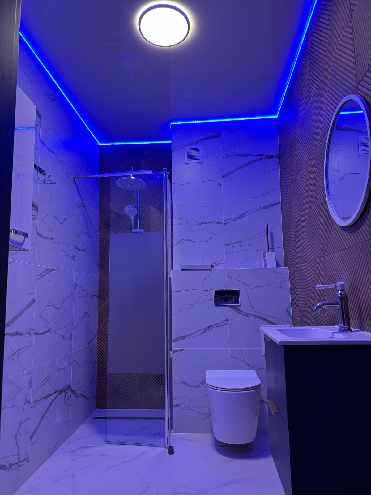 Nowoczesna łazienka z kabiną prysznicową typu walk-in, białymi płytkami imitującymi marmur, podświetleniem LED w kolorze niebieskim oraz okrągłym lustrem.