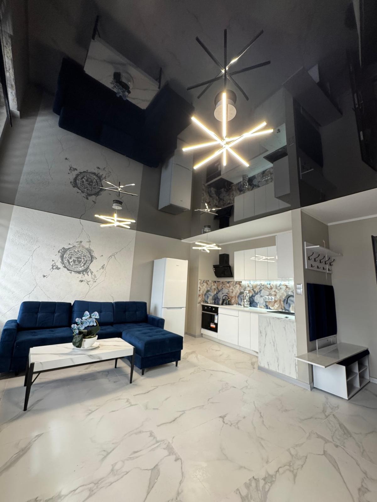 Salon z aneksem kuchennym w nowoczesnym stylu: błyszczący sufit, marmurowa podłoga, granatowy narożnik i białe meble kuchenne z dekoracyjnym panelem nad blatem.