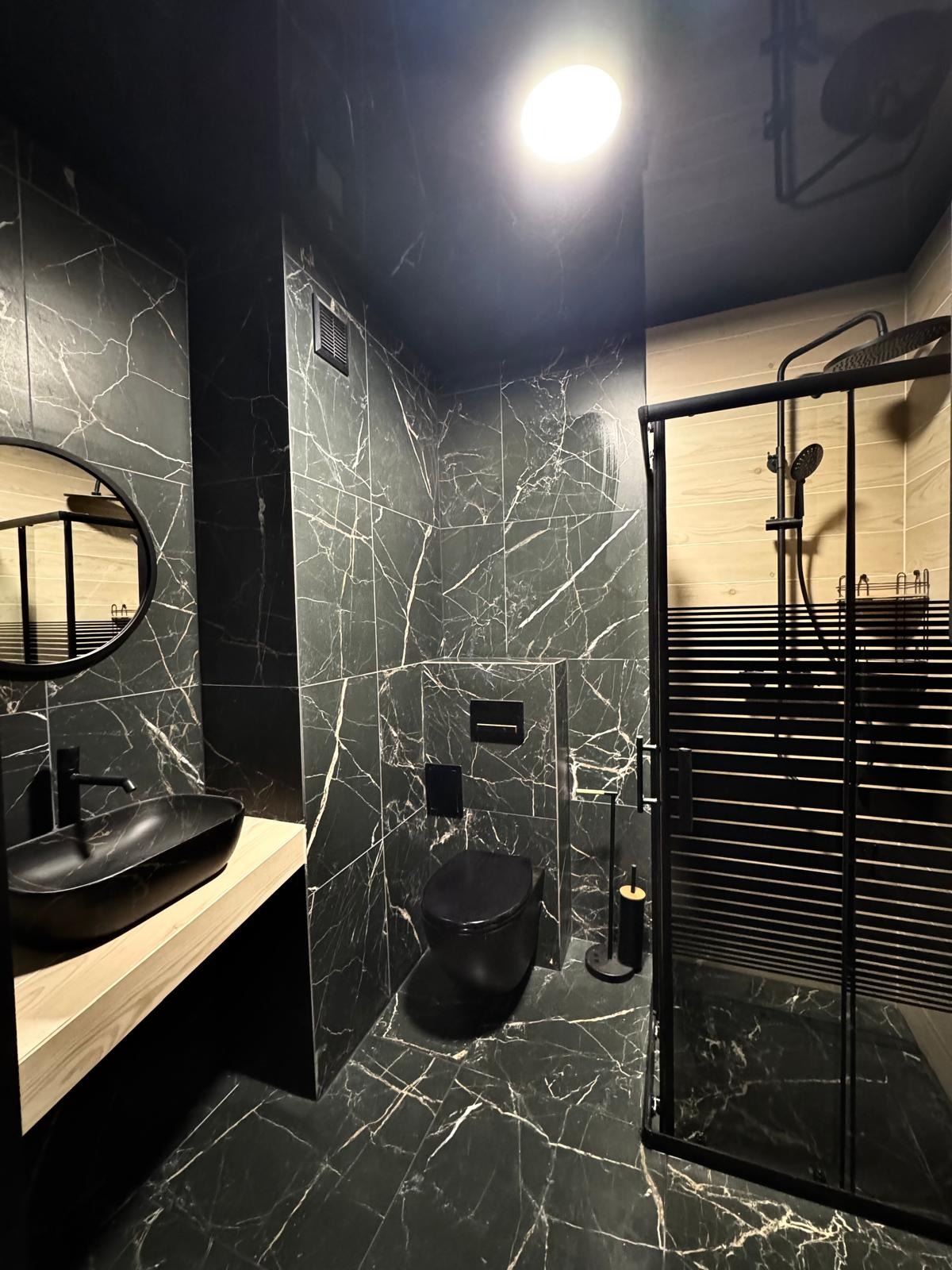 Nowoczesna łazienka z czarnymi płytkami imitującymi marmur, czarną armaturą i kabiną prysznicową typu walk-in z elementami drewna.