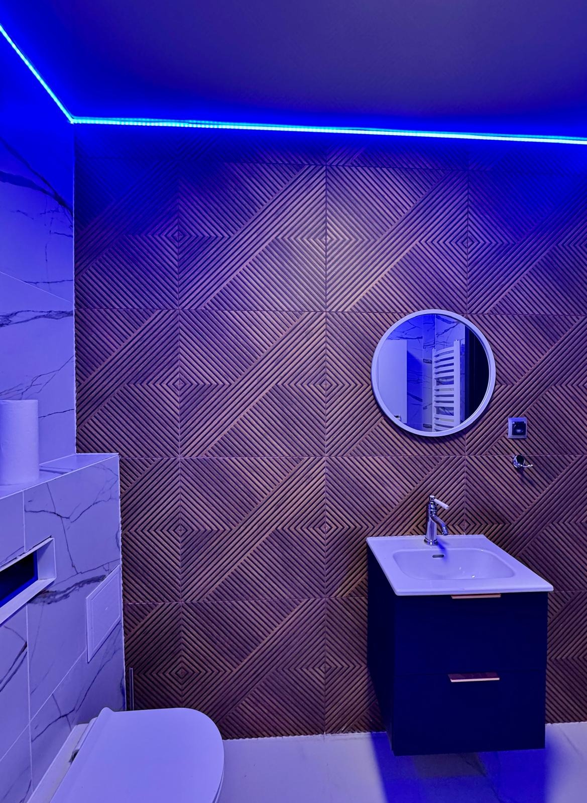 Nowoczesna łazienka z geometryczną ścianą w odcieniach brązu, podświetlona niebieskim światłem LED, okrągłe lustro nad umywalką z szafką w kolorze granatowym.