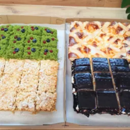 Dwa rodzaje ciast w kartonowych pudełkach: jedno z zieloną posypką i owocami, drugie z polewą i ciemną czekoladą, widok z góry.