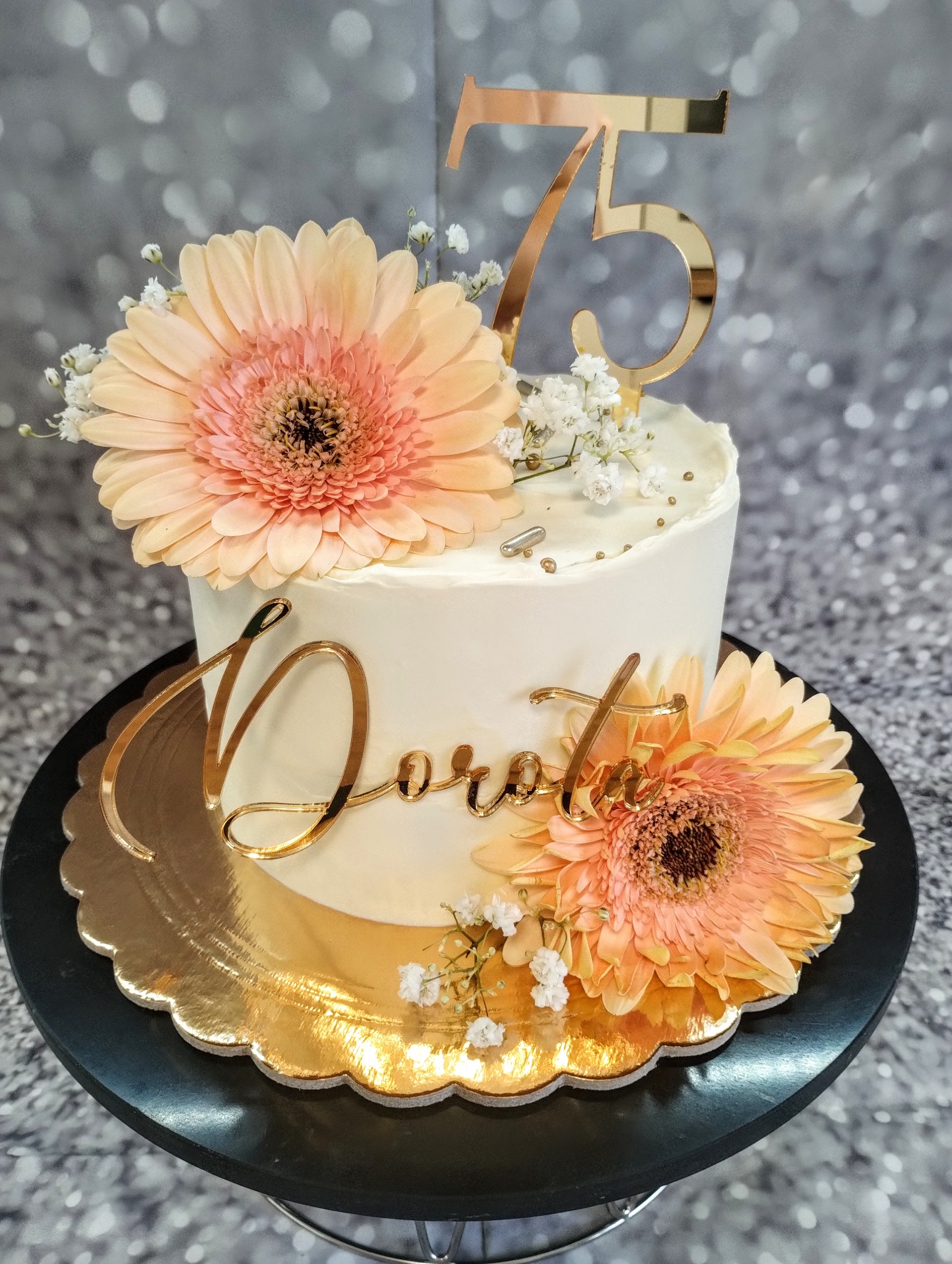 Tort urodzinowy z białym kremem, ozdobiony brzoskwiniowymi gerberami, gipsówką i złotym napisem 'Dorota' oraz numerem '75', umieszczony na złotej paterze.