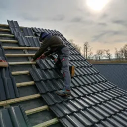 Dekarz w szarych spodniach roboczych układa ciemnoszarą dachówkę na drewnianej konstrukcji dachu w słoneczny dzień.