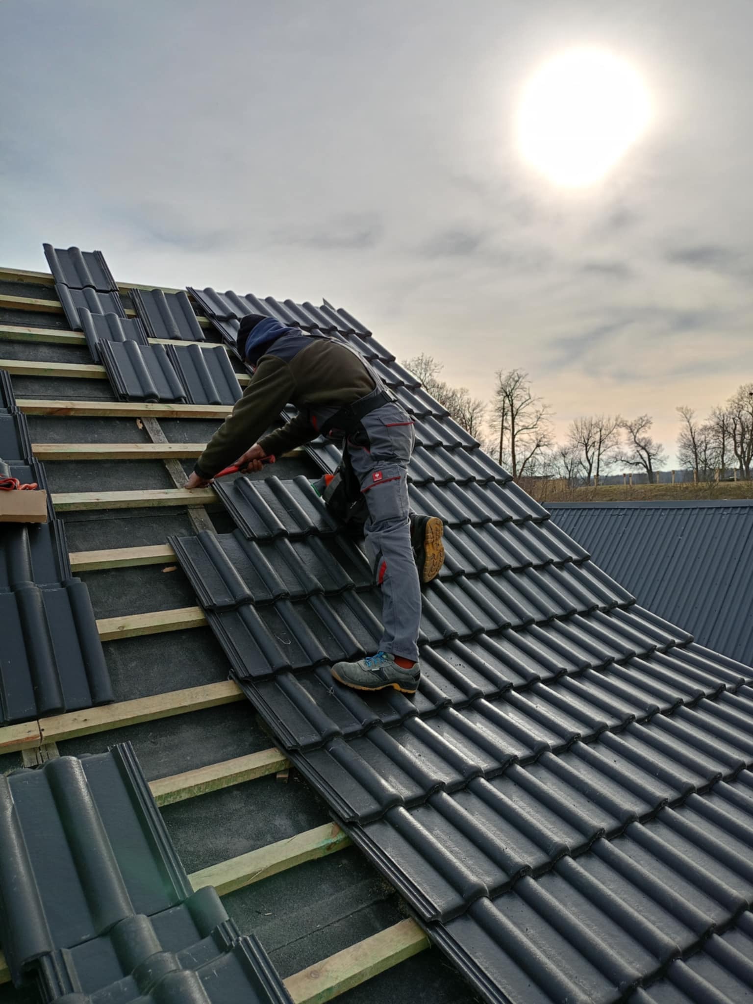 Dekarz w szarych spodniach roboczych układa ciemnoszarą dachówkę na drewnianej konstrukcji dachu w słoneczny dzień.