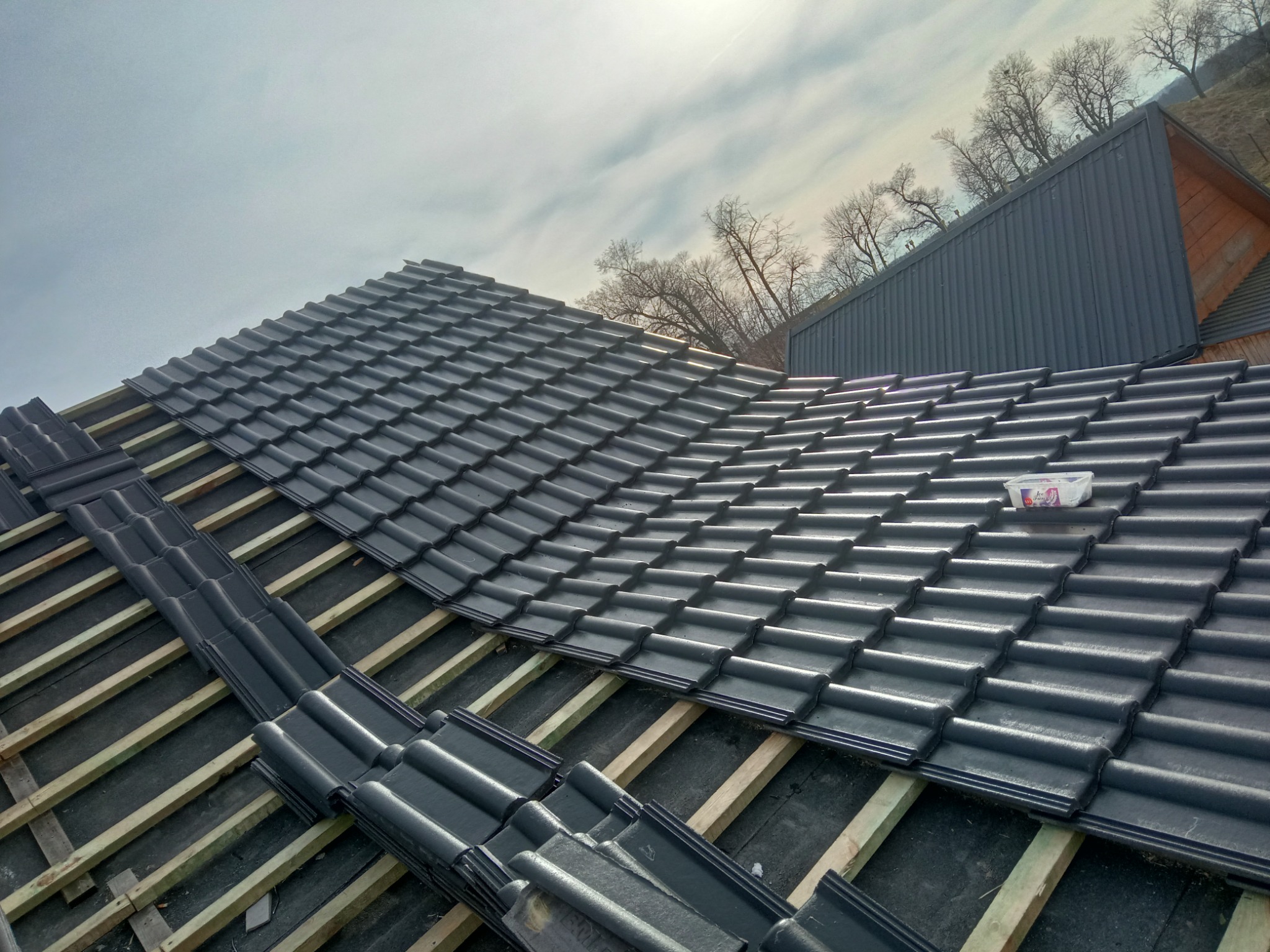 Montaż ciemnej dachówki ceramicznej na drewnianej konstrukcji dachu, widoczne niezakończone rzędy i pojemnik z tworzywa sztucznego na gotowej części dachu.