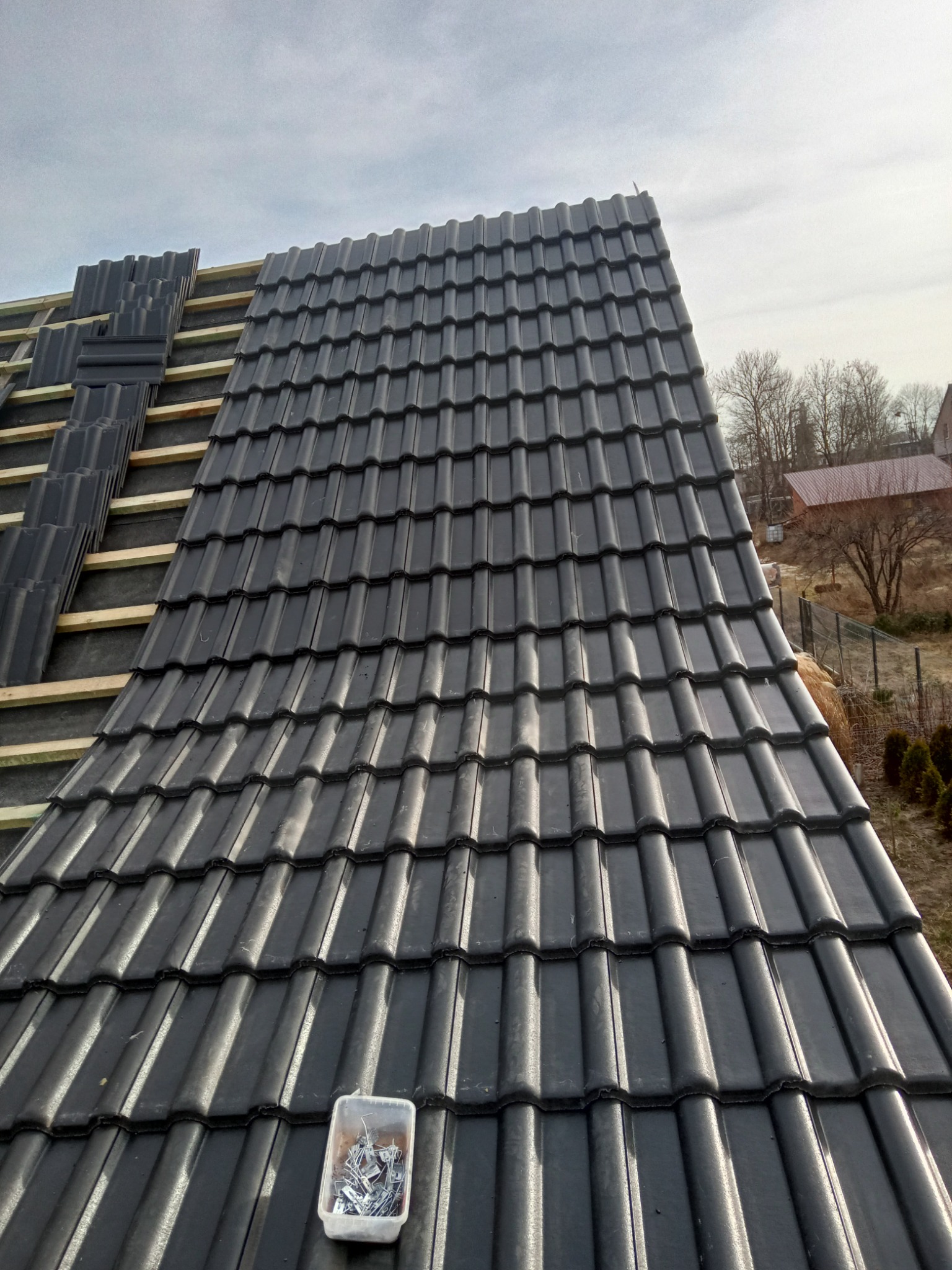 Fragment dachu w trakcie układania z widocznymi łatami, częściowo pokryty czarną dachówką, z pojemnikiem na metalowe klamry w dolnym rogu.