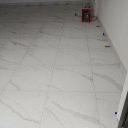 Szwagier usługi remontowo ,wykonczenowe - Wykończona podłoga z białych płytek imitujących marmur, z niebieskimi krzyżykami dystansowymi przy ścianach i grzejnikiem elektrycznym w rogu pomieszczenia.