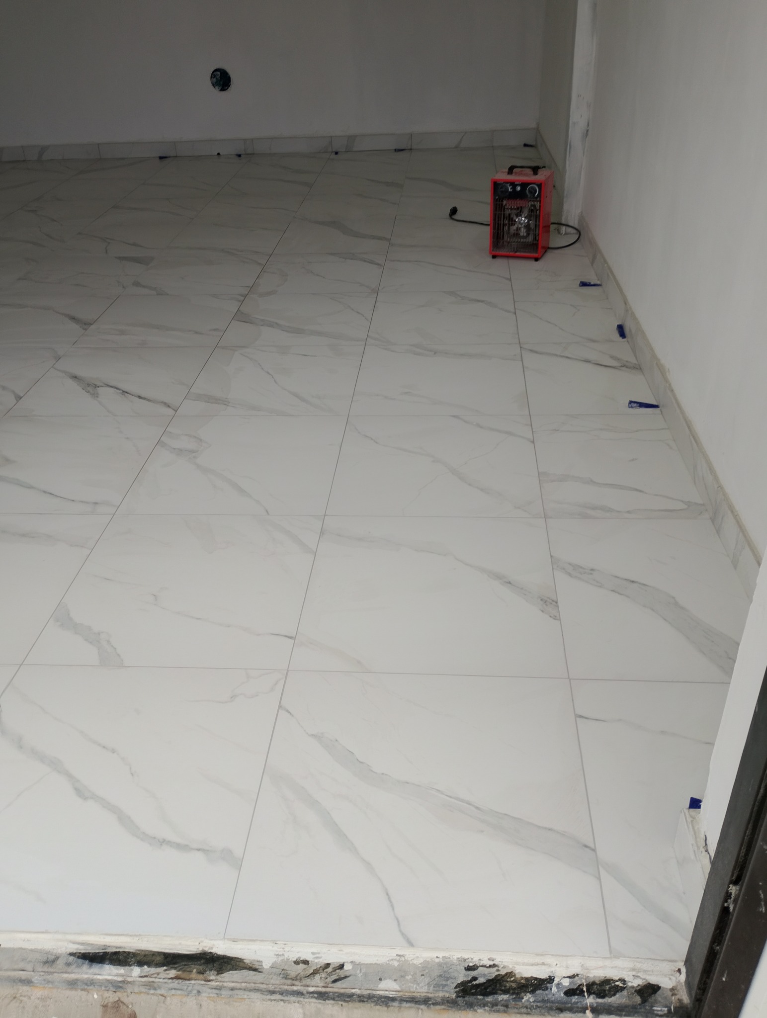 Wykończona podłoga z białych płytek imitujących marmur, z niebieskimi krzyżykami dystansowymi przy ścianach i grzejnikiem elektrycznym w rogu pomieszczenia.